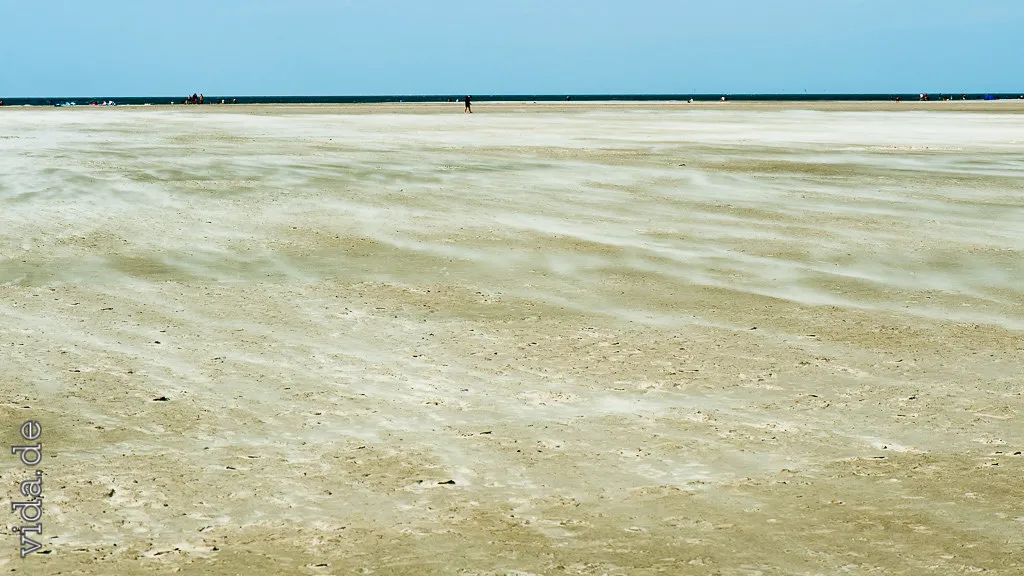 Weiter Sandstrand unter blauem Himmel, der Wind treibt den Sand in Schleiern