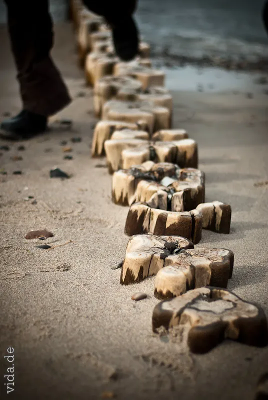 Holzklötze in einer Reihe im Sand, ein Fuß tritt ins Bild